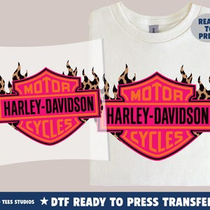 May include: A white t-shirt and a separate transfer sheet both featuring a pink and orange Harley-Davidson Motor Cycles shield logo with a black leopard print flame design. A blue circular badge in the corner displays the text Ready To Press.