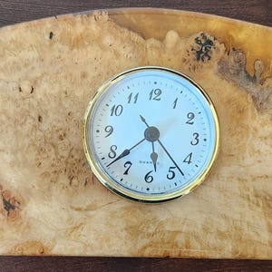 Op de afbeelding: Een handgemaakte houten bureauklok met een lichtblauwe wijzerplaat en goudkleurige rand. De klok is geplaatst in een unieke, gebogen houten basis met een natuurlijk houtnerfpatroon en een gouden harsrand. De klok is ongeveer 20 cm breed.