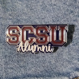 May include: A rectangular pin with the text "SCSU Alumni" in a maroon and silver color scheme. The pin is set against a blue denim background. The letters "SCSU" are in a bold, outlined font, and "Alumni" is in a script font.
