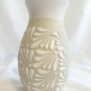 May include: A ceramic vase with a cream-colored body and a white top. The body features a raised, repeating white floral pattern. The vase has a rounded shape and a slightly flared opening, suitable for displaying flowers or as a decorative piece.