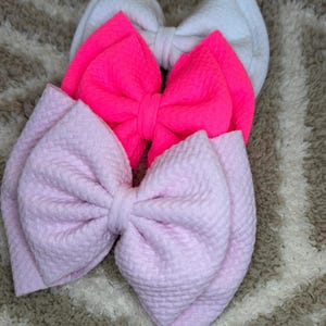 May include: Three fabric hair bows in a stack. The top bow is white, the middle bow is bright pink, and the bottom bow is light pink. The bows have a textured, quilted appearance. The background is a neutral, patterned surface.