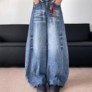Front view of a person wearing very baggy, wide-leg blue denim jeans with a vintage wash, featuring black shoes and a decorative red and black cord on the waistband.