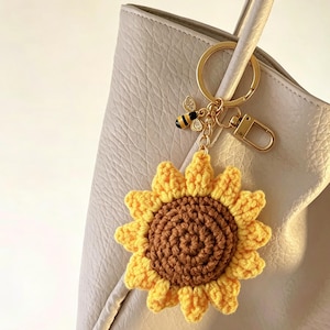 May include: A crochet sunflower keychain featuring a brown center and yellow petals, attached to a gold-toned metal ring and lobster clasp. A small, detailed bee charm with black and yellow stripes and clear accents hangs from the gold chain.
