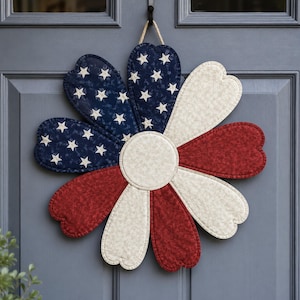 May include: A fabric flower wall hanging featuring heart-shaped petals in a patriotic color scheme. The petals are arranged in a circular pattern with navy blue fabric with white stars, solid red fabric, and off-white fabric with a subtle pattern.