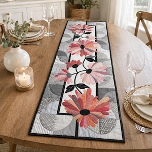 May include: A floral table runner with a black border. The runner features large, stylized flowers in shades of pink and orange, with black centers and stems. The background is a patchwork of gray and white geometric patterns.