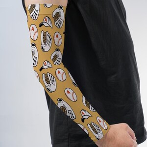 May include: An arm sleeve featuring a mustard yellow background with a repeating pattern of white baseball gloves, white baseball caps, and white baseballs with a red letter A printed on the front of each cap and baseball.