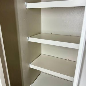 Könnte beinhalten: Ein Set aus fünf weißen, horizontalen Regalböden, die in einer Wandnische montiert sind. Jeder Regalboden ist mit kleinen weißen Metallwinkeln an der Seitenwand befestigt, was eine gestufte Aufbewahrungsmöglichkeit schafft.