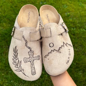 May include: A pair of beige suede slip-on clogs by Nautica featuring a buckle strap. The left shoe displays a black ink illustration of a leafy branch and a cross on a chain. The right shoe features a mountain range, a sun, and the text Psalm 121:1-2.
