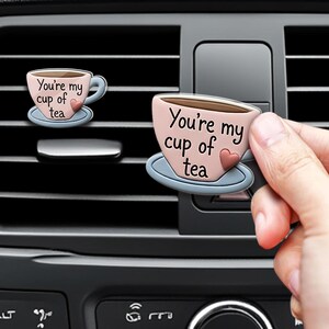 May include: A pink tea cup shaped car air freshener clip with a blue saucer and a small red heart. The text "You're my cup of tea" is printed in black on the front of the cup. The item is designed to attach to a vehicle's air vent slats.