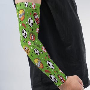 May include: A green arm sleeve featuring a repeating pattern of black and white soccer balls, red and yellow comic-style speech bubbles with the word GOAL, and small white stars scattered across the fabric, worn on an arm.