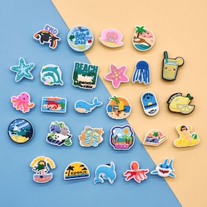 May include: A collection of colorful beach-themed charms, featuring images of sea creatures, tropical drinks, and beach scenes. Some charms have text that reads "END", "SAVE OUR SEAS", and "BEACH". The charms are made of a glossy material.