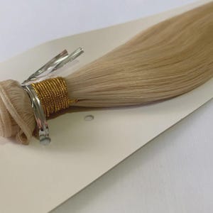May include: A bundle of light blonde hair extensions, secured with a metal clip and wrapped with gold-colored thread. The hair is smooth and appears to be human hair. The extensions are placed on a white card.