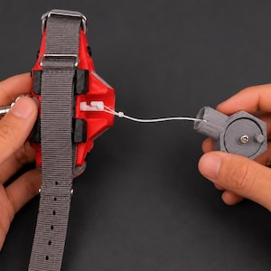 May include: A red and black device with a grey strap and a separate grey hexagonal component, connected by a white string. The device has a metallic silver clasp and is held against a dark background.