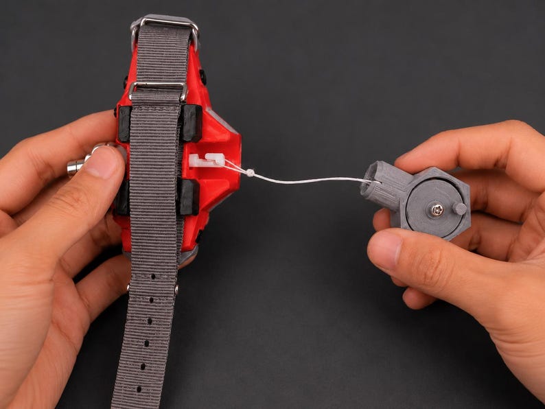 May include: A red and black device with a grey strap and a separate grey hexagonal component, connected by a white string. The device has a metallic silver clasp and is held against a dark background.