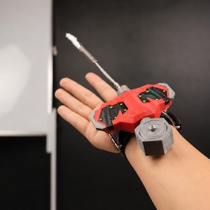 May include: A red and black wrist-mounted device with a silver and white extension. The device has a black strap and a grey hexagonal component. The device is designed to be worn on the wrist.