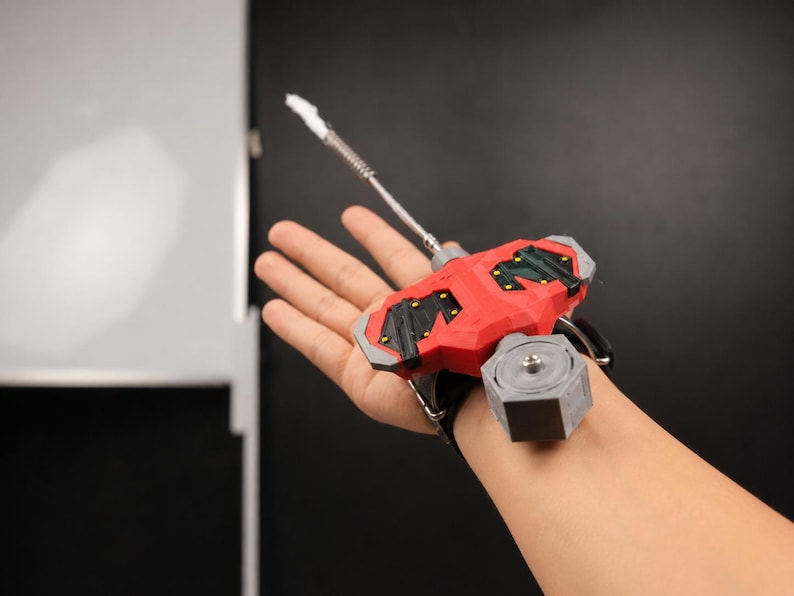 May include: A red and black wrist-mounted device with a silver and white extension. The device has a black strap and a grey hexagonal component. The device is designed to be worn on the wrist.
