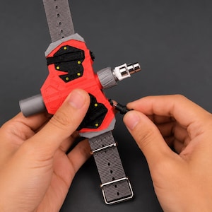 May include: A red and black device with a grey strap and a silver connector. The device has a complex design with multiple components and is being held by a person. The strap has a buckle and several holes.