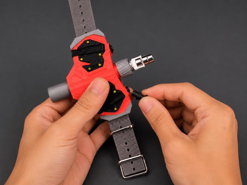 May include: A red and black device with a grey strap and a silver connector. The device has a complex design with multiple components and is being held by a person. The strap has a buckle and several holes.