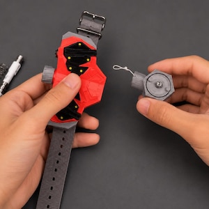 May include: A red and black wrist-worn device with a grey strap and buckle is held in one hand, while a detached grey component is held in the other. A white pen-like object and a silver spring are also visible. The device appears to be a prop or a toy.