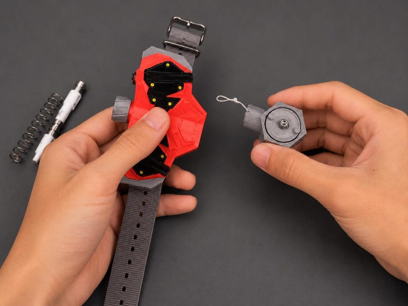 May include: A red and black wrist-worn device with a grey strap and buckle is held in one hand, while a detached grey component is held in the other. A white pen-like object and a silver spring are also visible. The device appears to be a prop or a toy.