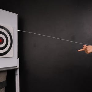 May include: A white target with black concentric circles and a red centre is hit by a thin, white string. A device on a wrist shoots the string. The target is on a white board, and the background is black.