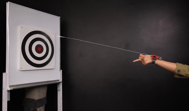 May include: A white target with black concentric circles and a red centre is hit by a thin, white string. A device on a wrist shoots the string. The target is on a white board, and the background is black.