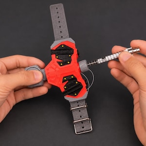 May include: A red and black wrist-mounted device with a grey strap and buckle. The device has a cylindrical component with a spring and a silver-coloured tip. The device is held by two hands.