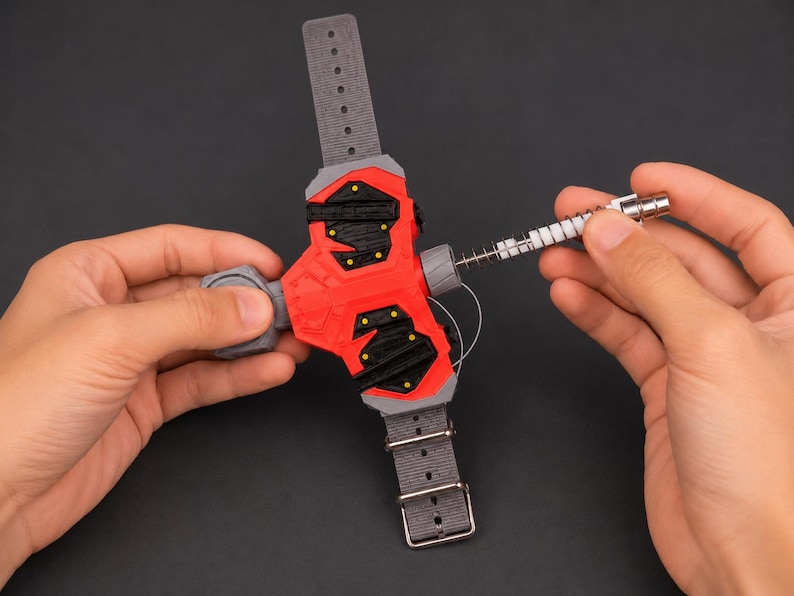 May include: A red and black wrist-mounted device with a grey strap and buckle. The device has a cylindrical component with a spring and a silver-coloured tip. The device is held by two hands.