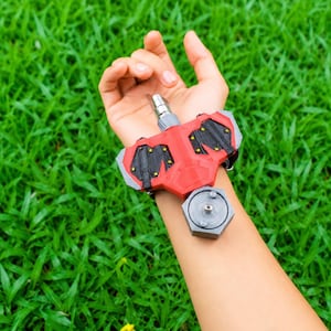 May include: A red and grey wrist-mounted device with black accents and yellow details. The device has a cylindrical nozzle and a hexagonal component. The device is worn on a wrist against a backdrop of green grass.