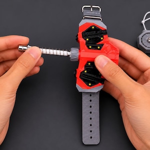 May include: A red and grey wrist-mounted toy with a spring-loaded mechanism, featuring a grey strap and a separate grey disc with a white string. The toy is held by hands against a dark background.