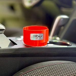 May include: A bright red cup holder with the text "Cup Corral" and a bull graphic on a white label. The cup holder is in a car's center console, designed to fit in a standard cup holder space.