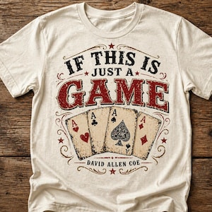 May include: Cream-colored t-shirt featuring the text "IF THIS IS JUST A GAME" in a distressed, vintage style. Below the text are four playing cards, each displaying an Ace. The shirt also includes the text "DAVID ALLEN COE".