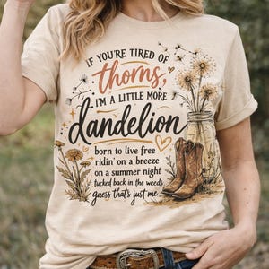 Può includere: T-shirt beige con la scritta "If you're tired of thorns, I'm a little more dandelion". La maglietta presenta una grafica di dente di leone, stivali da cowboy e un barattolo di vetro. La persona indossa jeans blu e una cintura marrone.