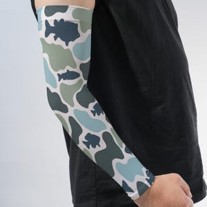 May include: An arm sleeve featuring a camouflage-inspired pattern in shades of light blue, sage green, and dark charcoal. The design incorporates silhouettes of various fish scattered throughout the irregular, organic shapes of the print.