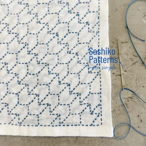 Op de afbeelding: Close-up van een witte stof met een geometrisch patroon gestikt met blauw garen. Het ontwerp heeft een herhalend patroon van onderling verbonden vormen. De tekst "Sashiko Patterns" is zichtbaar. Een naald en draad zijn ook aanwezig.