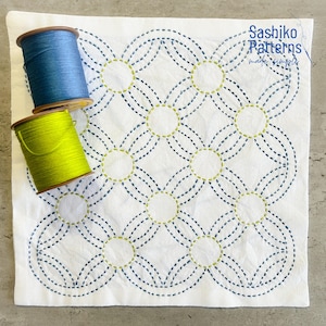 May include: A white fabric square featuring a repeating geometric pattern of interlocking circles stitched in dark blue and lime green thread. Two spools of thread, one blue and one lime green, rest on the fabric. Text reads Sashiko Patterns.