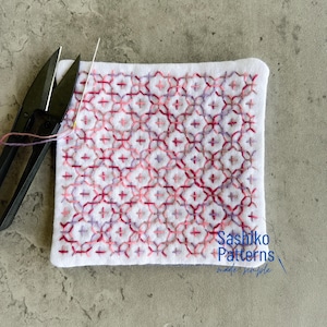 May include: White square fabric with a geometric pattern embroidered in shades of red and purple. A needle and thread are in the fabric, with a pair of black scissors to the left. The text "Sashiko Patterns made simple" is in the bottom right corner.