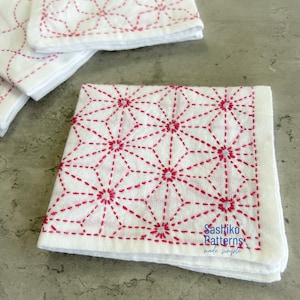 May include: White cloth napkins with a red geometric stitched pattern. The napkins are folded and stacked, showcasing the detailed embroidery. The text "Sashiko Patterns made simple" is visible on one napkin.