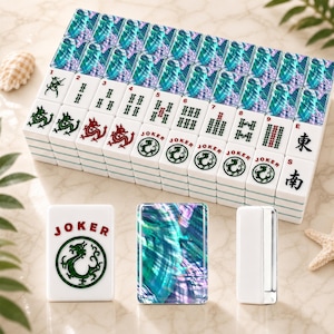 May include: A set of white Mahjong tiles featuring iridescent blue and green abalone shell backs. The tiles display various red and green engraved symbols, including dragons, numbers one through nine, Joker tiles, and the East and South wind characters.