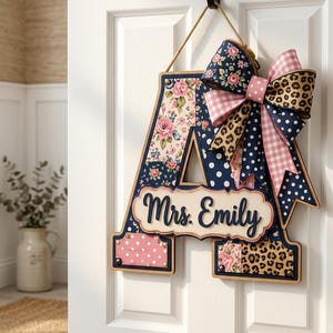 May include: A decorative letter A door hanger featuring a patchwork design of floral, polka dot, and leopard print patterns. A large multi-patterned bow is attached to the top right, and a central plaque displays the text Mrs. Emily in script.