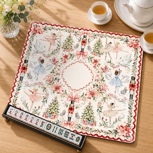 May include: A square mahjong mat featuring a cream background with a festive pattern of nutcrackers, ballerinas, swans, Christmas trees, and poinsettias, bordered by a scalloped red edge, accompanied by a rack of white mahjong tiles.