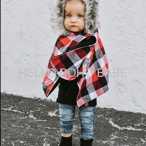May include: A child wearing a fur-lined hood and a red, black, and white plaid cape. The child is also wearing a black top, distressed blue jeans, and black boots. The text "HELLO BOHO BABE" is visible on the image.