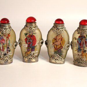 May include: Four antique Chinese snuff bottles. Each bottle features a red stopper, silver-toned metal filigree, and a painted scene. The bottles have a light yellow background and are displayed on a white surface.