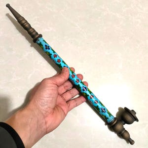 May include: A long, slender smoking pipe featuring a bright turquoise shaft decorated with a colorful floral pattern. The pipe has a metallic, bronze-toned mouthpiece at one end and a matching bronze-toned bowl at the opposite end.