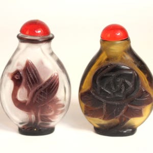May include: Two small glass snuff bottles with rounded bodies and red spherical stoppers. One bottle is clear with a carved dark purple bird design, while the other is amber-coloured with a carved dark brown rose flower motif on the front.