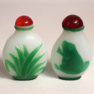 Puede incluir: Dos frascos de rapé de vidrio blanco opaco con cuerpos redondeados y tapones de vidrio rojo. Un frasco presenta un relieve de vidrio verde de una planta de hojas, mientras que el otro muestra un relieve de vidrio verde de una rana sentada.