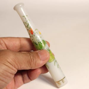 May include: A cylindrical white translucent cigarette holder featuring a hand-painted floral design with green leaves, orange and yellow petals, and a thin gold band near the base. The object has a smooth, polished finish and a flared mouthpiece.