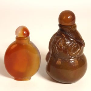 May include: Two small snuff bottles made of polished brown and orange agate. The bottle on the left is round and smooth, while the bottle on the right has a gourd-like shape with carved leaf details and a matching rounded stopper on top.
