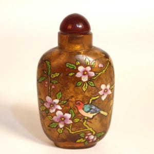 Puede incluir: Una pequeña botella de vidrio marrón antiguo con un tapón rojo oscuro. La botella está decorada con un pájaro colorido posado en una rama con flores rosas y hojas verdes. La botella tiene forma redondeada.