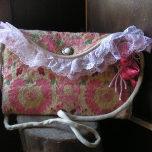 May include: A vintage-style handbag with a floral pattern and lace trim. The bag has a pink and white lace trim and a pink ribbon accent. The bag is made of a fabric with a floral pattern.